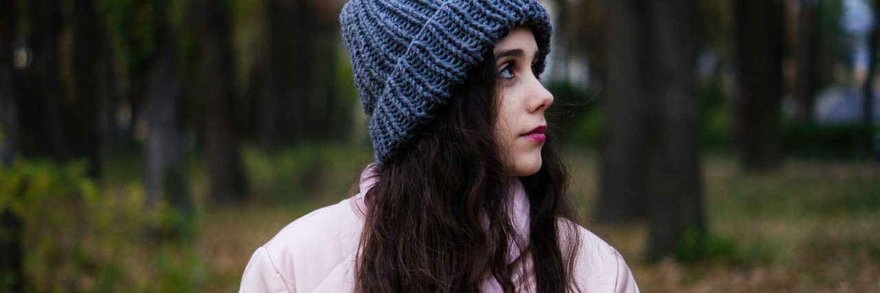 Having a Chronic Illness Isn't Your Fault young woman with long dark hair wearing a pink jacket and blue hat standing outside in the woods