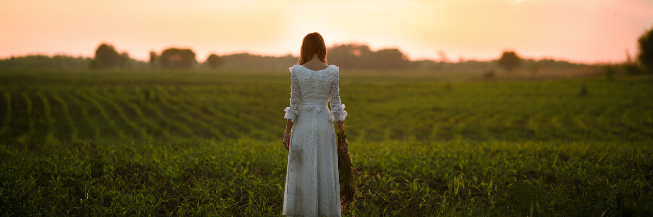 Young woman in long white lace dress on cornfield at sunset. Back view.