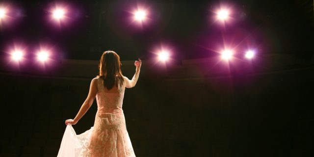 A woman in a gown on a stage with stage lights focusing on her.