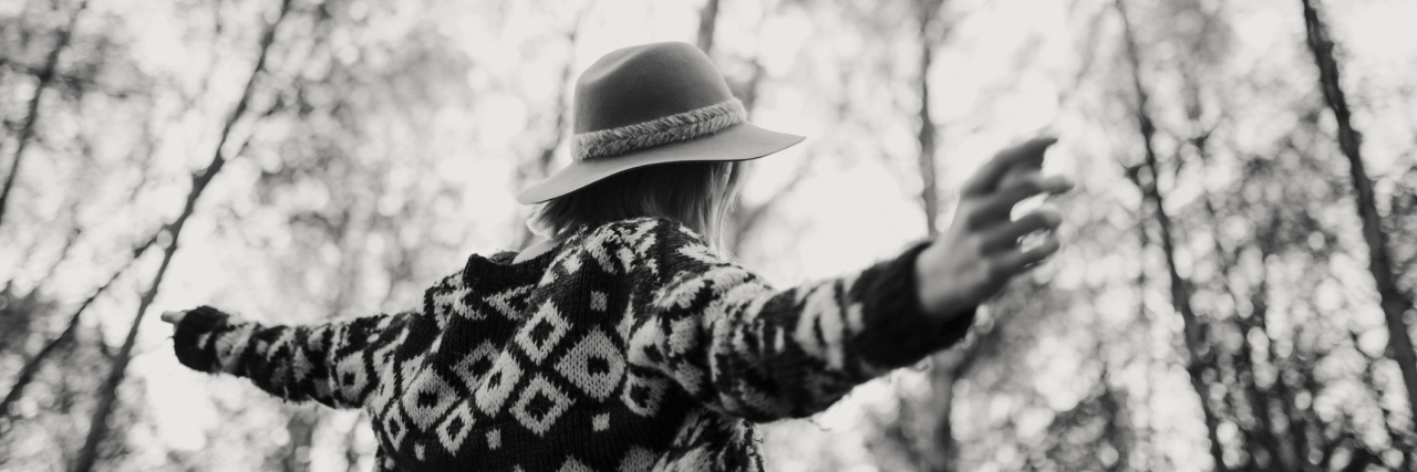 black and white photo of a woman wearing a sweater and a hat while walking in the forest