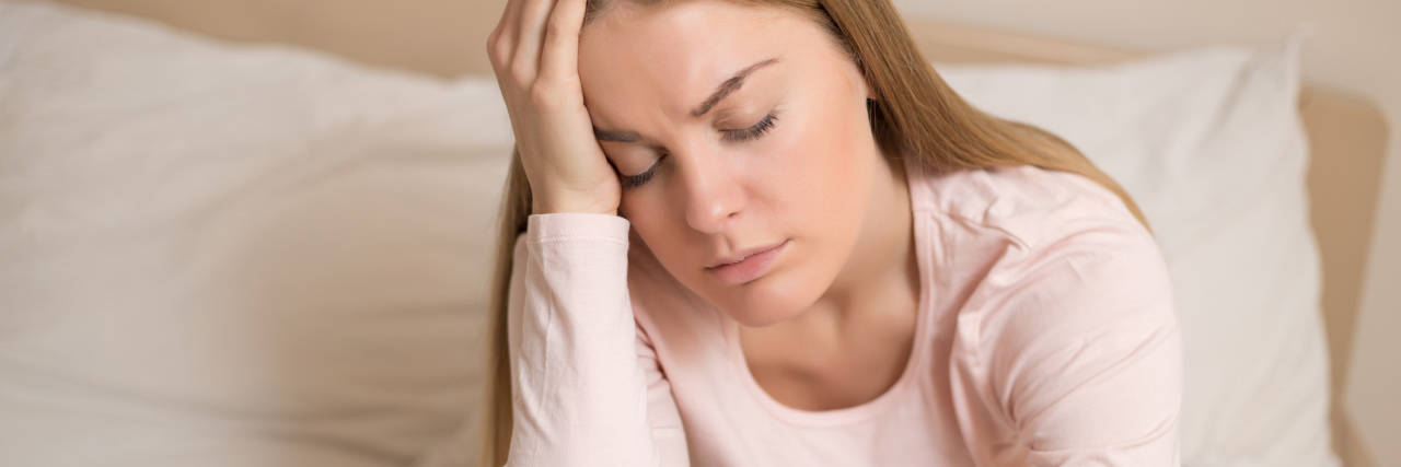 Struggling With Basic Tasks When You're Chronically Ill woman sitting on her bed resting her head against her hand with her eyes closed