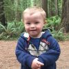Little boy with Down syndrome, woods in background, he is smiling, holding his hads together and wearing a blue sweatshirt.