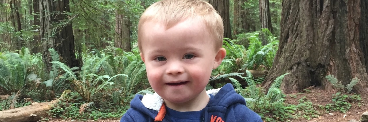 Little boy with Down syndrome, woods in background, he is smiling, holding his hads together and wearing a blue sweatshirt.