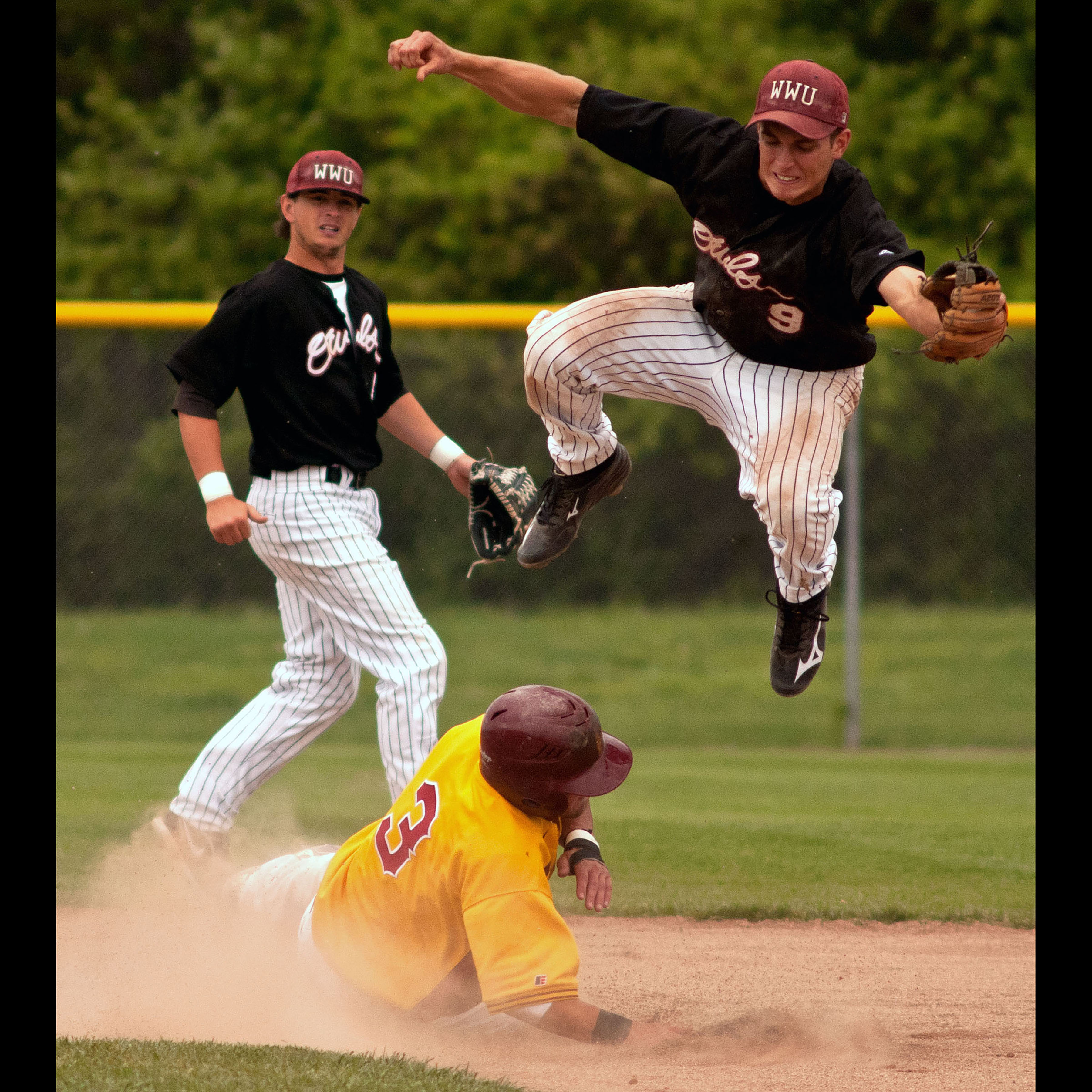 A photo of a baseball player in the air trying to catch a ball, while a player from another team slides into base.