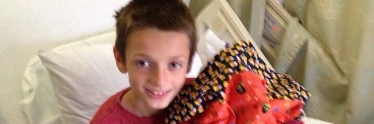 When My Son Was Diagnosed With Type 1 Diabetes Boy sitting on side of hospital bed wearing a red sweatshirt, and holding a dark orange pillow