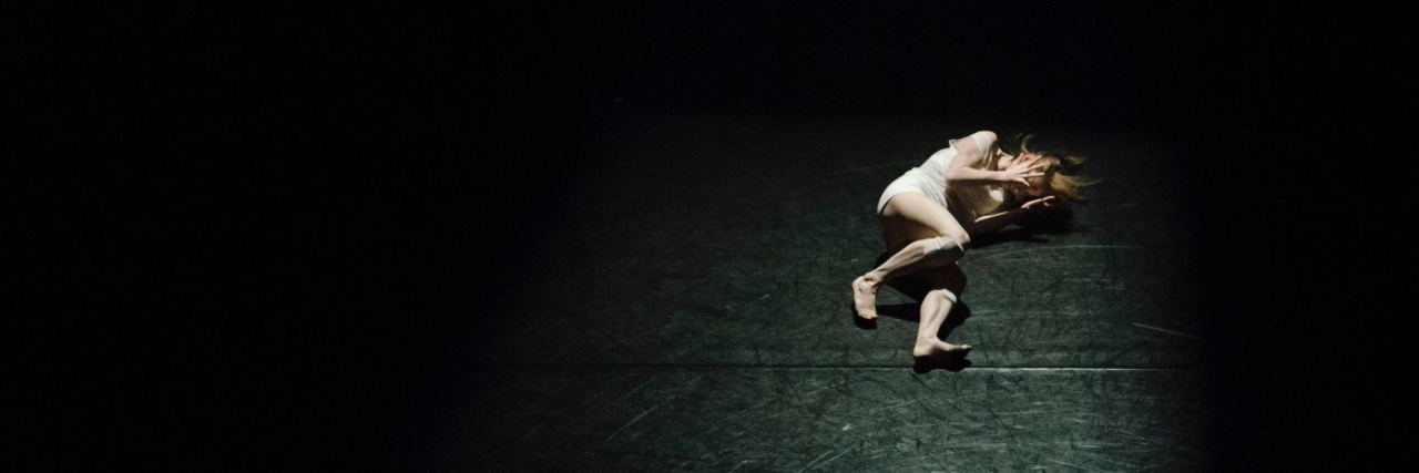 11 Photos That Can Help Explain Chronic Illness woman lying on a dark floor illuminated by a spotlight