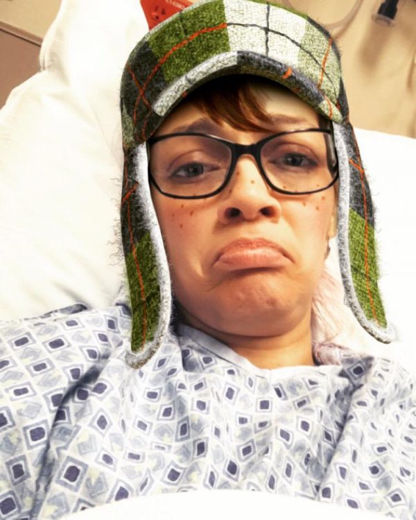 woman wearing glasses and a warm hat while lying in a hospital bed and frowning