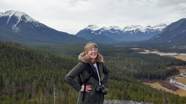 woman wearing a puffy jacket with a camera around her neck smiling with her hands on her hips at the summit of a mountain overlooking a mountain range and forest in alberta