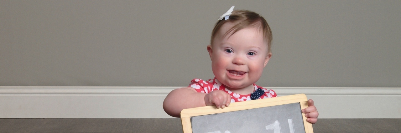 To the Mom Told to Abort Your Baby With Down Syndrome Little girl with Down syndrome sitting on floor and holding a sign that reads, "I'm 1!"