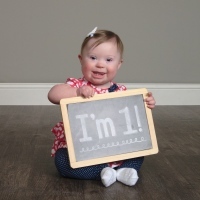 Little girl with Down syndrome sitting on floor and holding a sign that reads, "I'm 1!"
