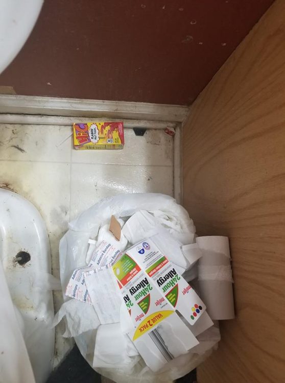 the corner of a bathroom with a basket of trash and a juicebox lying on the floor