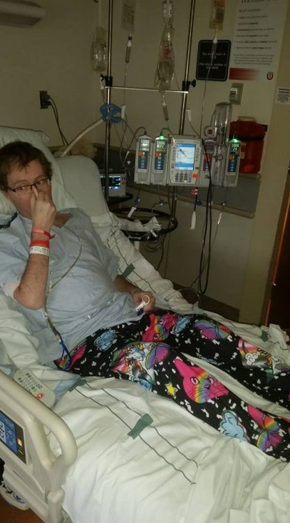 man in a hospital bed covering his nasal tube with his hand and wearing pajama pants with rainbow-colored kittens on them