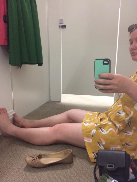 woman in a yellow dress sitting on the floor of a changing room in a store and taking a selfie in the mirror