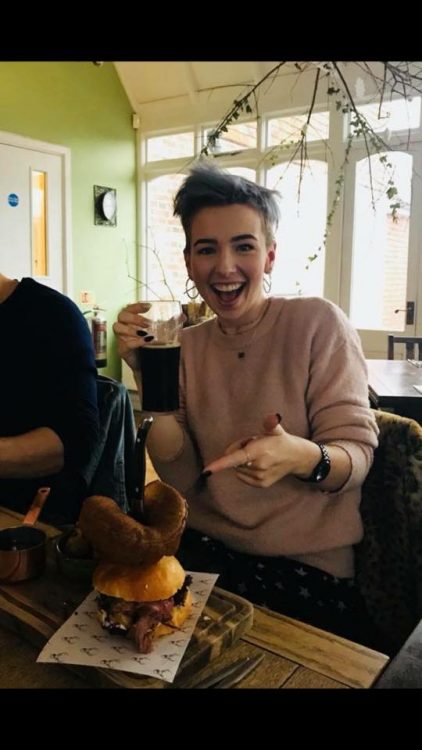 woman with short blue hair sitting in a restaurant smiling and pointing at a burger