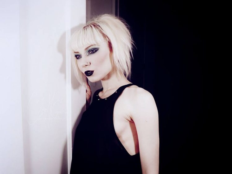 photo of a woman with blonde hair wearing a black shirt and purple lipstick and leaning against the wall in a pose