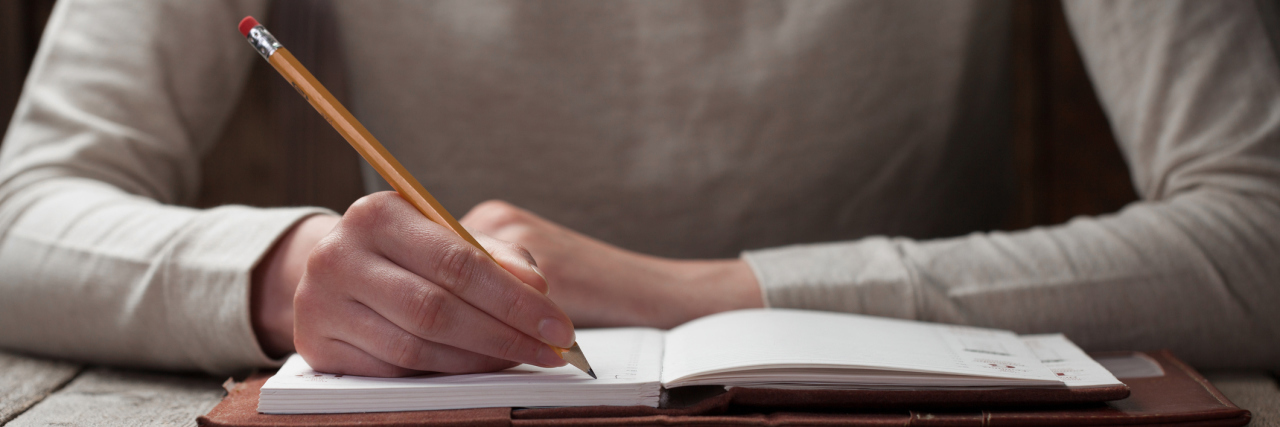 How Writing Has Helped Me Cope With My Chronic Pain A woman writing with a pencil in a journal.