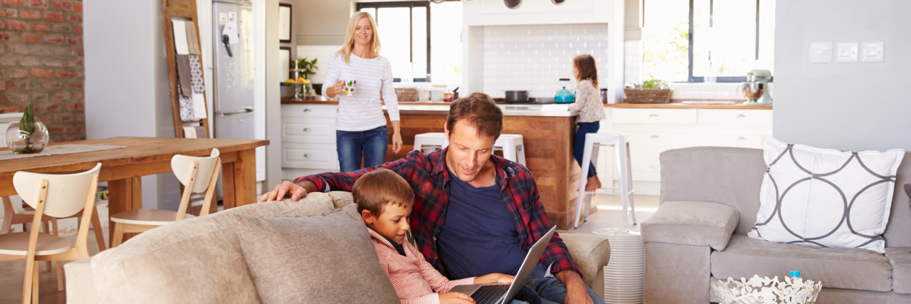 Embracing My New Life After Retiring Early Due to Multiple Sclerosis a man helping his son work on a computer in their living room with the mother and daughter in the kitchen