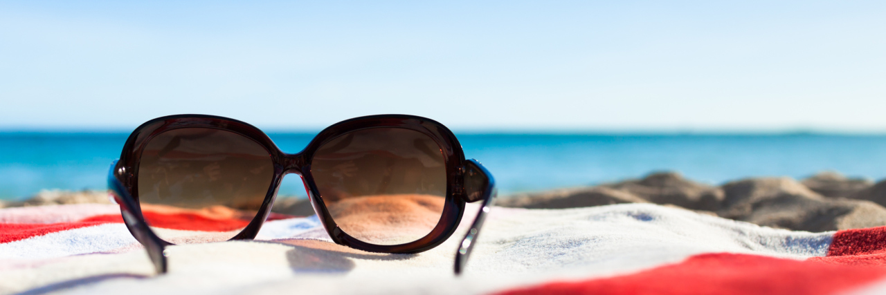 What Spring Break Is Like for Someone With Chronic Illness sunglasses sitting on a red and white striped towel on the beach
