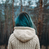 woman with blue hair wearing a white sweater and standing in front of a pine forest