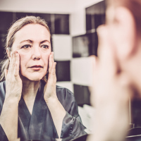 A woman touching her face and looking in the mirror at her reflection.
