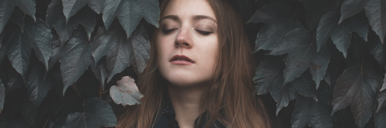 Being Honest About Illness Is Different Than Being Negative woman with brown hair standing with her eyes closed against a wall of leaves