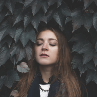 woman with brown hair standing with her eyes closed against a wall of leaves