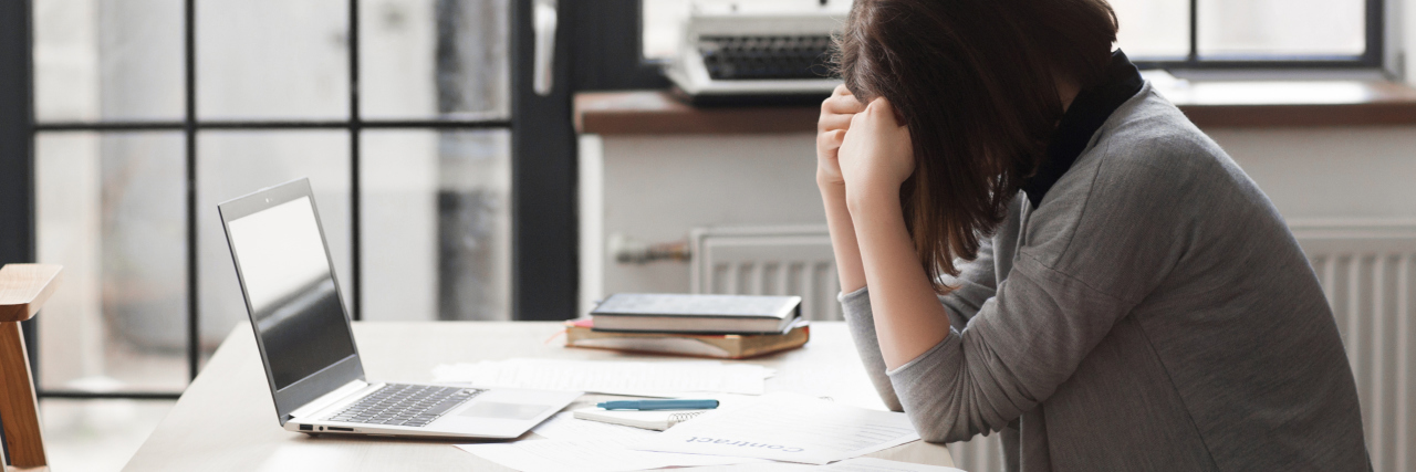 Feeling 'Lazy' When You Can't Work Due to Chronic Illness woman sitting at a desk in an office holding her head in her hands