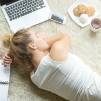 A college student laying down and napping while surrounded by her books, computer, a snack, coffee, and her phone.