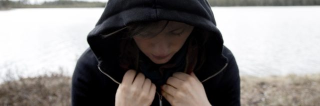 Woman in a black hoodie standing with the back towards a lake. She is looking down and has a shadow over her face. Concept photo of depression, mental illness and loneliness.