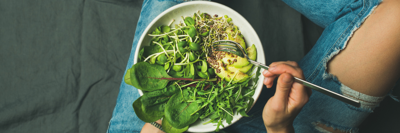 How the Vegan Lifestyle Impacted My Eating Disorder Recovery Green vegan breakfast meal in bowl with spinach, arugula, avocado, seeds and sprouts. Girl in jeans holding fork with knees and hands visible, top view, copy space. Clean eating, vegan food concept