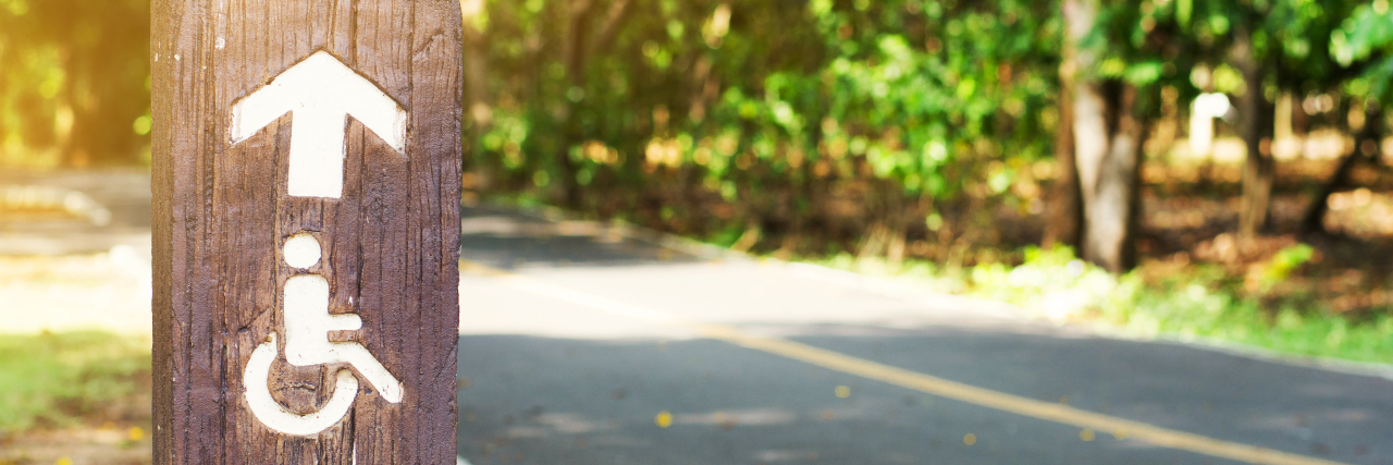 Can Disability Be an Identity Without Defining Us? Disability sign with arrow pointing towards trail.