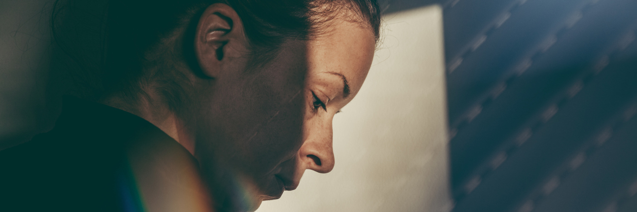 Why I've Had to Trust My Gut During My Journey With Chronic Illness A woman leaning against a wall, partially covered by a shadow, looking upset.
