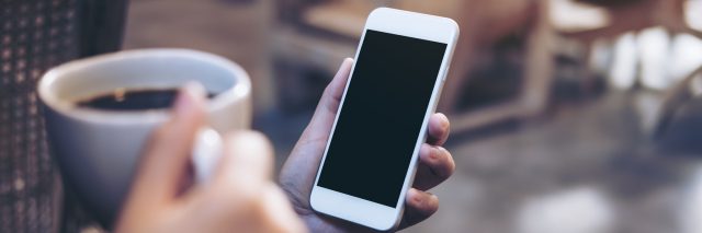 Mockup image of woman's hands holding white mobile phone with blank black screen while drinking coffee in modern loft cafe