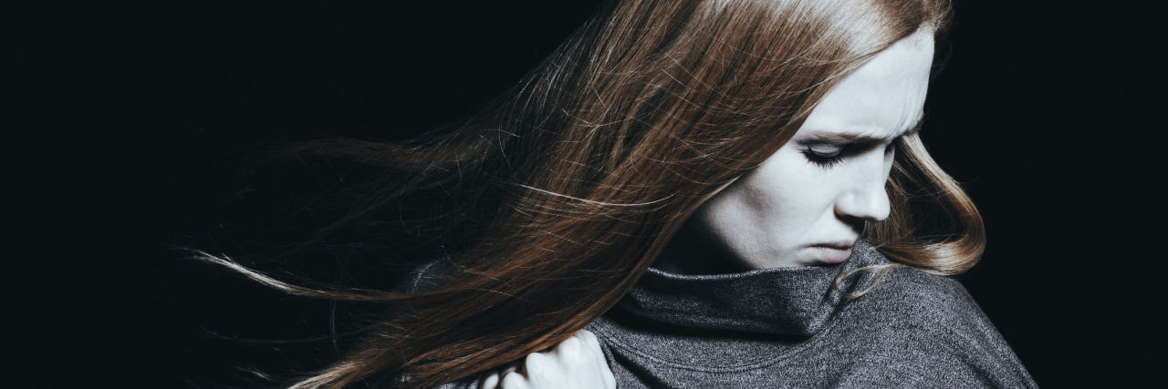 How Our Culture Is Still Hungover From Diagnosing Women With Hysteria A woman in a dark room, arms crossed, as she looks down.