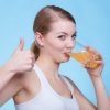 Food, health concept. Woman holding glass of orange flavored drink and drinking from it showing thumb up gesture. Studio shot on blue background