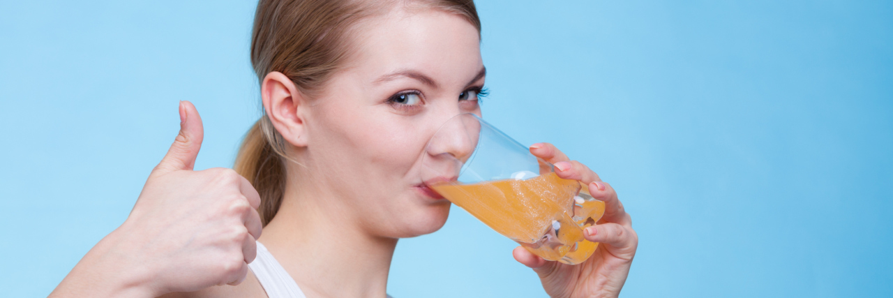 10 Cures for Chronic Illness You'll Find on the Internet Food, health concept. Woman holding glass of orange flavored drink and drinking from it showing thumb up gesture. Studio shot on blue background