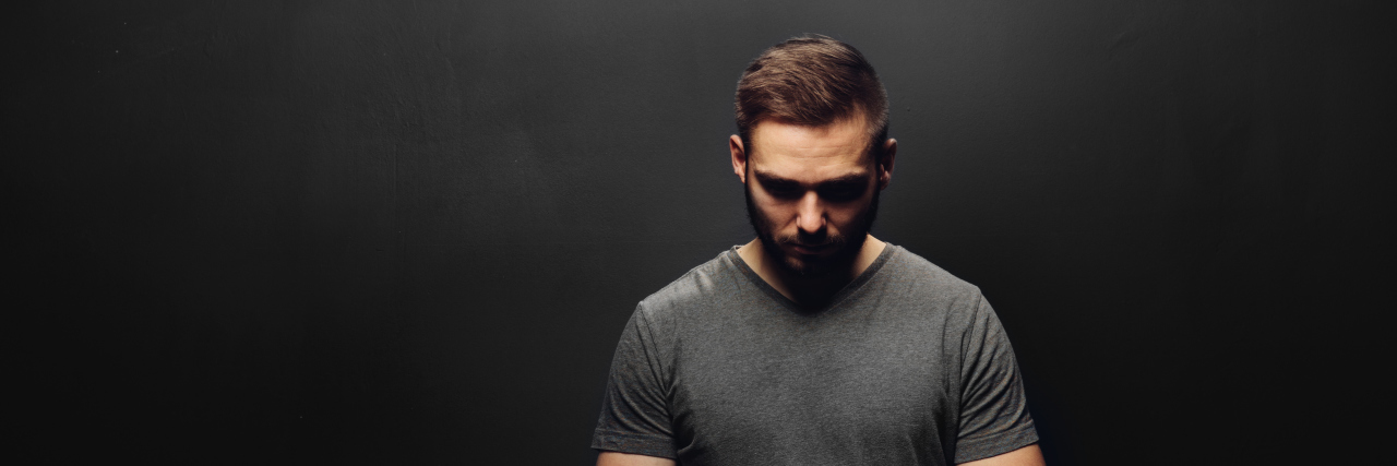 Why I Will Never Own a Gun as Someone With Suicidal Thoughts Young man standing on a black background, looking down in a mysterious pose.