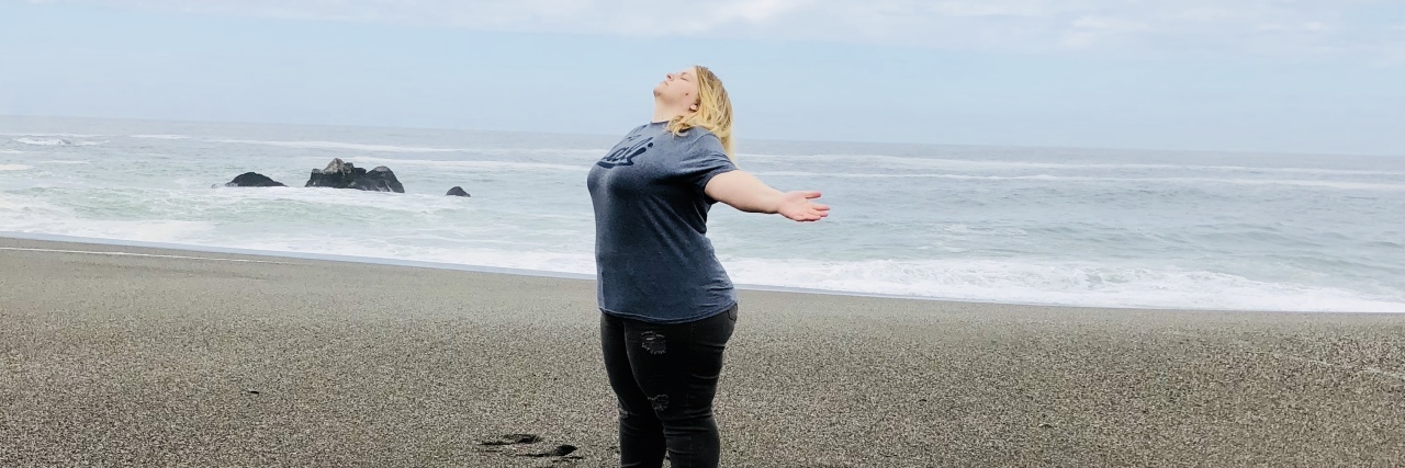 You Shouldn't Need to Apologize for Having an Illness woman standing on the beach and spreading her arms and looking up to the sky