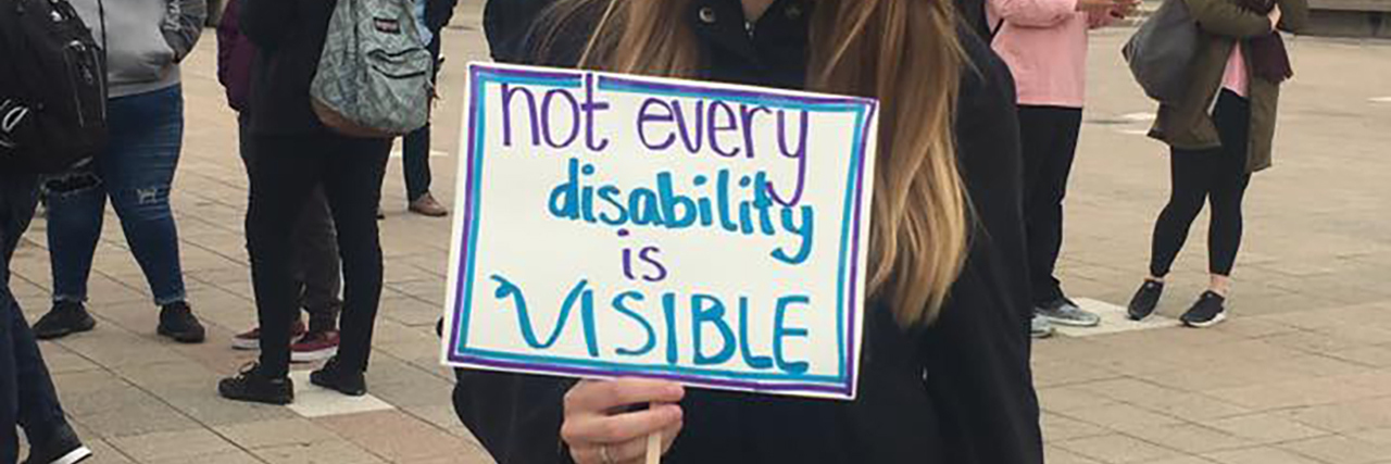 Zoie stands in the center of the UIC quad, pavement under her feet. In the background is one building and several individuals engaged in conversations. She is wearing a black jacket, her blonde hair is just beyond her shoulders, and she is white. In her right hand, she holds a white sign with blue border that says “not every disability is visible.”
