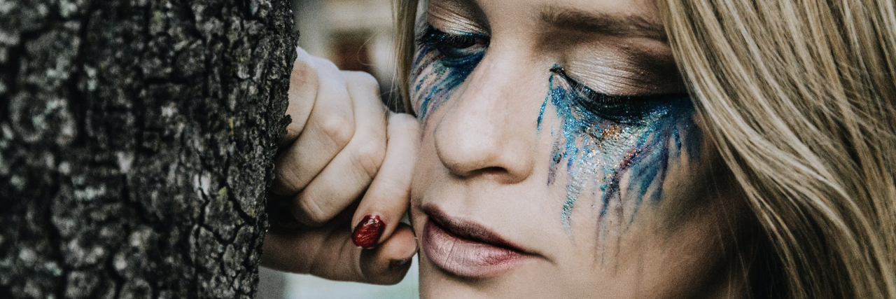 The Uncommon Migraine Symptoms I Experience close-up of a blonde woman leaning against a tree with turquoise makeup beneath her eyes