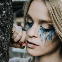 close-up of a blonde woman leaning against a tree with turquoise makeup beneath her eyes