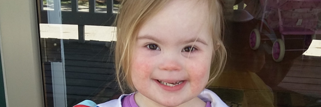 Little girl with Down syndrome wearing a snow white dress over her clothes and eating a bowl of cheese with a green spoon. She is smiling at the camera.