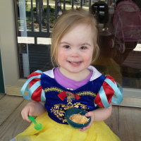 Little girl with Down syndrome wearing a snow white dress over her clothes and eating a bowl of cheese with a green spoon. She is smiling at the camera.