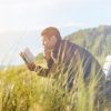Man sitting by sea on bench near tall grass reading a book