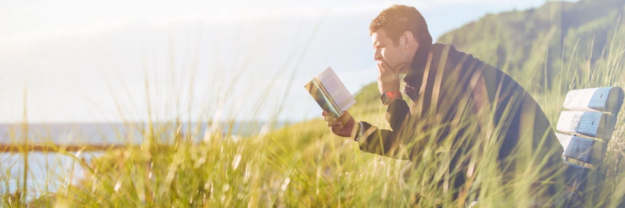 Man sitting by sea on bench near tall grass reading a book
