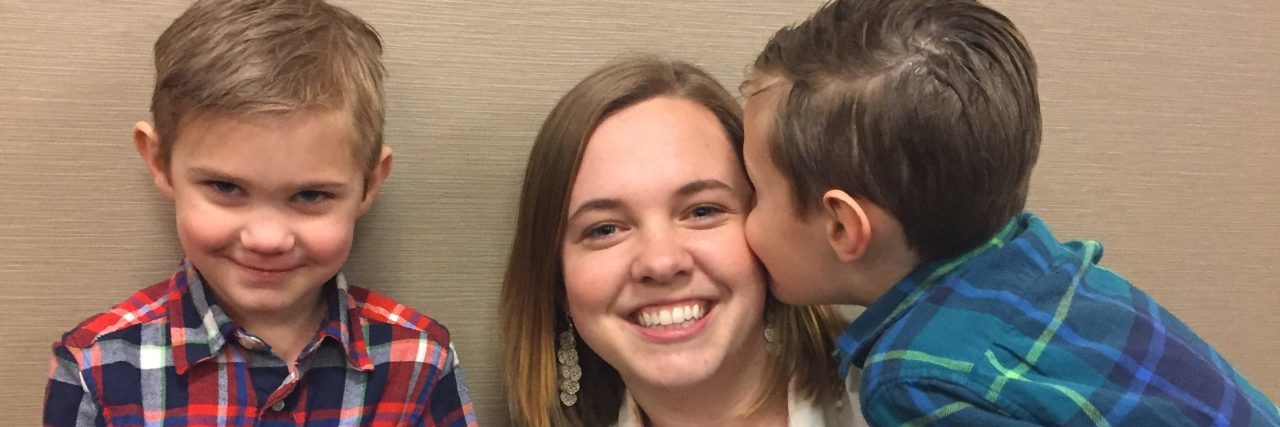 Having Children With Disabilities Transformed Me Mom holding a sign that reads: "Kissing booth" while one of hr son's kisses her on the cheek