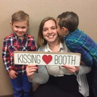 Mom holding a sign that reads: "Kissing booth" while one of hr son's kisses her on the cheek