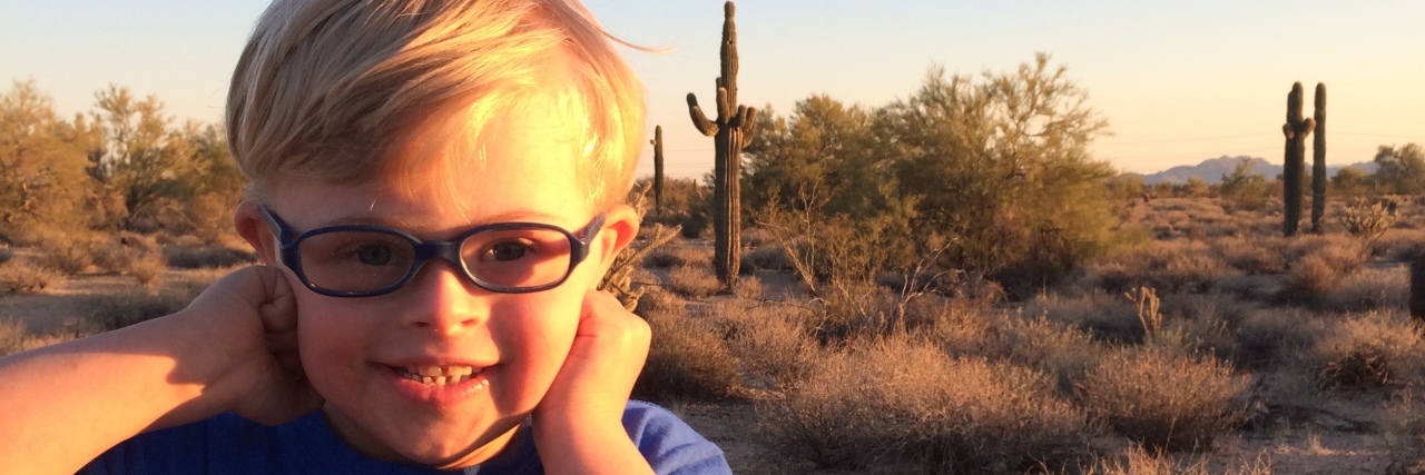 My Son With Down Syndrome Made Me a Believer Little boy with Down syndrome smiling. He wears a blue shirt, glasses and has blond hair.