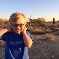 Little boy with Down syndrome smiling. He wears a blue shirt, glasses and has blond hair.