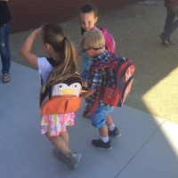Three children holding hands walking into school, in the middle is a little boy with Down syndrome